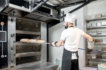 Baker taking off baked breads with shovel from the professional oven at the manufacturing - Powered by Adobe