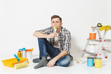 Smiling fun man sitting on floor with paint brush like mustache, instruments for renovation apartment isolated on white background. Wallpaper, gluing accessories, painting tools. Repair home concept.