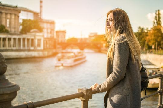 Side view of young woman looking at the sunset over a river in the city of Berlin with the old buildings and tourist boat in the background. 