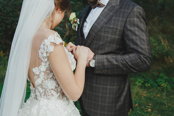 Beautiful wedding couple in the forest. The bride with tulle veil and open low back elegant dress is touching the groom in bow tie. Wedding buttonhole and checkered suit in Great Gatsby style.