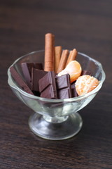 Chocolate with cinnamon sticks and tangerines in a transparent dessert plate on a dark table