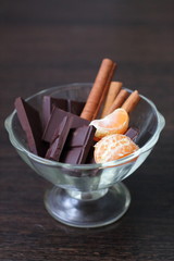 Chocolate with cinnamon sticks and tangerines in a transparent dessert plate on a dark table