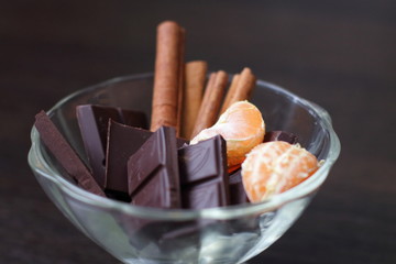 Chocolate with cinnamon sticks and tangerines in a transparent dessert plate on a dark table