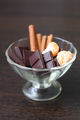 Chocolate with cinnamon sticks and tangerines in a transparent dessert plate on a dark table