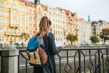 A young beautiful girl stands and looks at the map next to the Vltava River with the amazing old architecture of Prague in the background with a sunny spring day