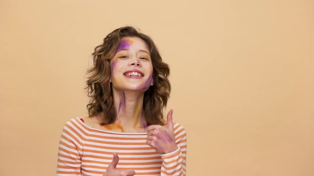 Portrait of smiling funny woman with multicolored art on her face suddenly appearing on camera and showing thumbs up slow motion, over beige background. Concept of emotions