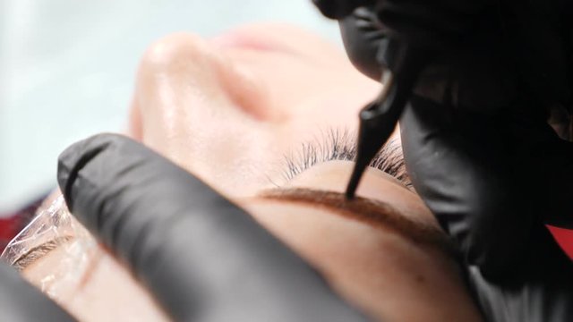 Beauty salon. Close up of Male beautician in black gloves making permanent makeup procedure on female eyebrows. Young woman gets facial beauty procedure. Facial rejuvenation. Using tattoo machine