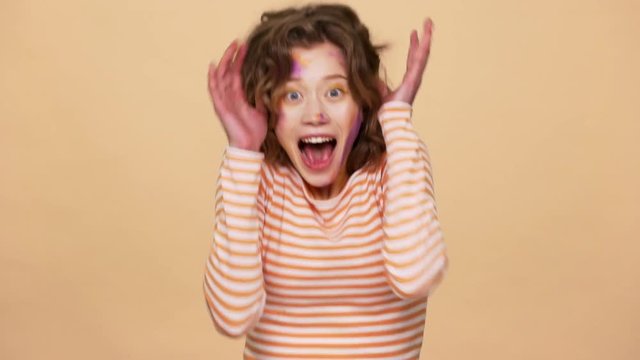 Portrait of ecstatic woman 20s european appearance with colorful stains on face screaming in happiness and delight slow motion, isolated over beige background. Concept of emotions