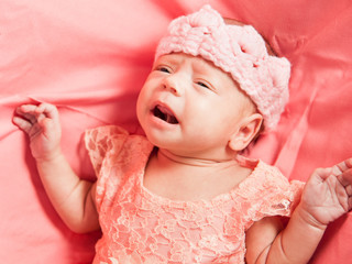 Newborn baby girl in pink dress and crochet crown, lies on pink bed.