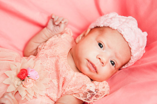 Newborn baby girl in pink dress and crochet crown, lies on pink bed.