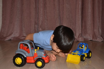 Little brown-eyed brunette boy in blue t-shirt plays on the floor with cars, tractor and bulldozer toys