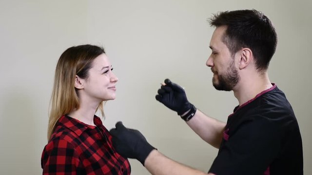 Portrait of happy female client and male beautician in healthcare clinic cabinet of beauty salon. Male cosmetologist in black gloves making permanent makeup procedure on female eyebrows. Thums up