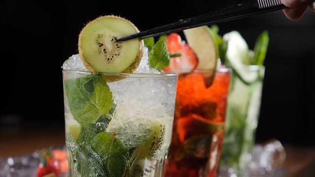 Final touch concept. Bar and cocktail concept. Bartender prepare different Mojito Cocktails. Close up. Adding a slice of qiwi on top of glass with cocktail. Slow motion.