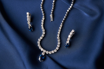 Women's platinum necklace and earrings with a diamond and blue precious sapphire stone on a silk blue background, close-up. Luxury female jewelry