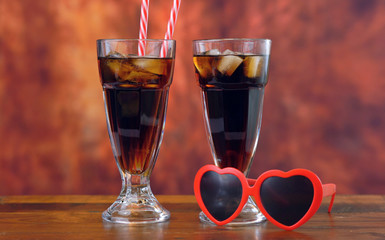 Pouring cola soft drink on ice in tall cafe glasses against a rustic wood background.