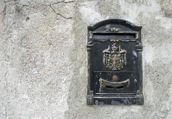 ancient letterbox, mailbox, black and gold, metal, set in wall outside house, in the center engraved a coat of arms (emblem) or banner, used by the postman to deliver letters, city concept, Italy
