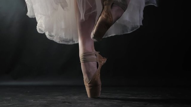 Close up of ballet dancer as she practices exercises on dark stage or studio. Woman's feet in pointe shoes. Ballerina shows classic ballet pas. Slow motion. Flare, gimbal shot.