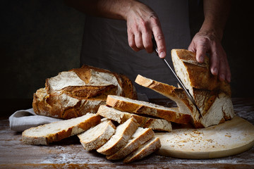 Hands cutting into slices a traditional loaf of freshly baked bread on wooden table in dark setting...