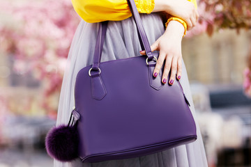 Close up photo of trendy violet bag with fur trinket in hands of fashionable woman posing in street...