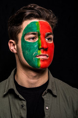 Portrait of handsome man face supporter fan of Portugal national team with painted flag face isolated on black background. Fans emotions.