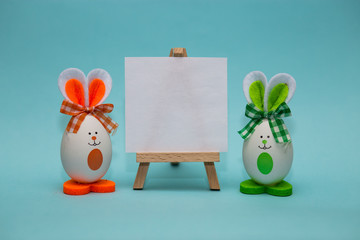 White blank paper on easel with funny eggs in form of cute bunny on blue background. Happy easter....