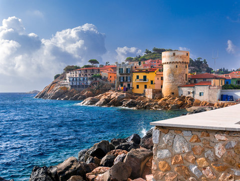 Perfect tiny seaside village of "Giglio Porto" with multi colored houses, an ancient defensive tower and a rocky coastline against a deep blue Mediterranean sea. - Giglio Island, Tuscany, Italy