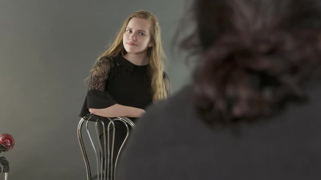Studio portrait photography. Female photographer works with blonde young model. Teen girl in black dress poses on grey background. Backstage shot during photoshoot.