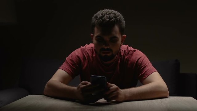 Depressed man sitting in dark empty room with smartphone, waiting for call