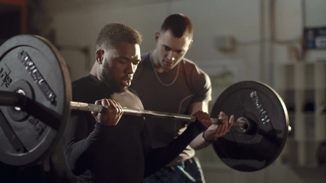 Concentrated African-American sportsman training bicep with heavy barbell under control of professional coach in gym.