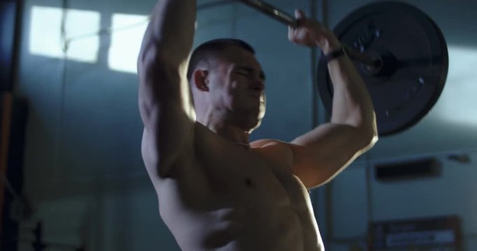 Concentrated athlete without shirt training in gym lifting heavy barbell above head training shoulders.