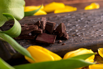 Pieces of dark chocolate on a decorative board surrounded by yellow tulips.