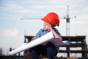 pensive handsome little kid in helmet solves problem of project on construction site with building crane and building in the background