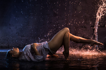 Girl in the white shirt with water drops and dark walls background illuminated by light during a...