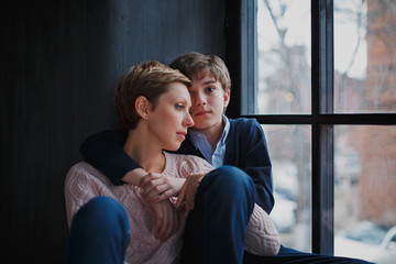 A teenage boy hugs his young mother with short hair and looks at the camera with sad eyes. The woman looks out the window.