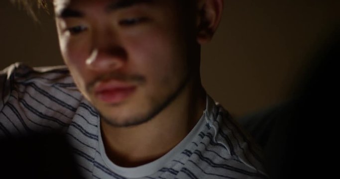 4K Close up on face of young Asian guy streaming media on digital tablet late at night