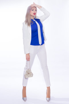 Studio portrait in full length of a beautiful young blond woman with long hair in a white pants suit. A girl in silvery shoes with high heels, in her hands she holds a small silver handbag