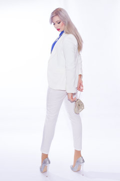 Studio portrait in full length of a beautiful young blond woman with long hair in a white pants suit. A girl in silvery shoes with high heels, in her hands she holds a small silver handbag