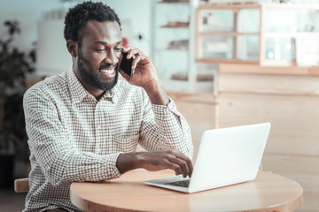 Business conversation. Positive cheerful nice man looking at the netbook screen and pressing a button while talking on the phone