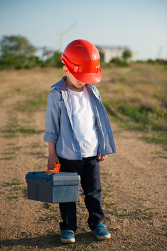 sad little working kid in orange helmet hung his head with tool case in one hand standing on path