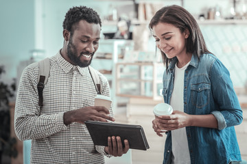 Digital device. Happy positive nice man holding a cup of coffee and showing a tablet to his friend while talking together with her
