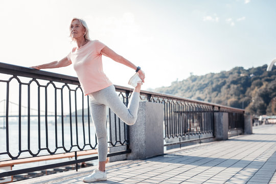 Forever young. Senior athletic pleasant woman standing on the quay holding by the railing and doing exercises.