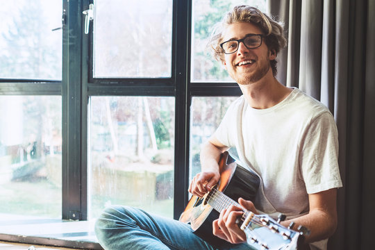 Happy smiling young man play on guitar near the window