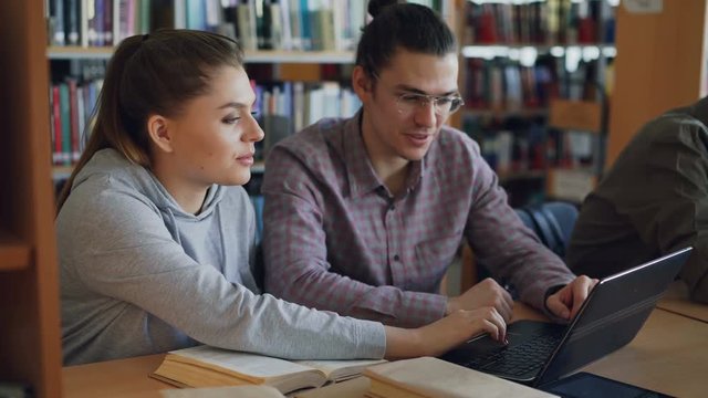 Cheerful female and male college students working on laptop together while sitting at table at university library with classmates on background