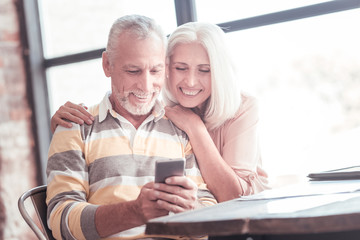 Show me. Satisfied cute aged couple sitting by the table smiling and looking at the smartphone.