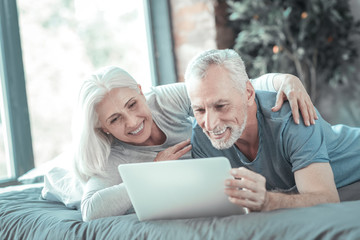 Moments of happiness. Joyful cute gray haired couple lying on the bed using the tablet and smiling.