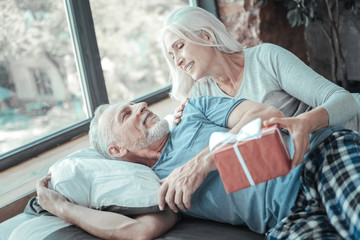 Unexpected gift. Cute pleasant senior woman lying on the bed near her husband smiling and giving the present.