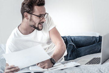 Fototapeta premium Working at home. Cheerful intelligent handsome man holding documents and looking at the laptop screen while being involved in work