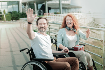 Hey you. Positive disabled woman keeping smile on her face and holding present on knees while holding hand on her boyfriend