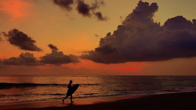 Silhouette of boy running along sea beach at sunset carrying surf board and enjoying beautiful sunset time on coast of Bali island in indonesia. Slow motion video
