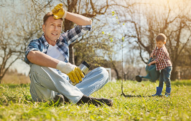 Make a pause. Tired pleasant adult man sitting on the grass while his little son watering a tree in the garden